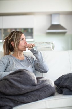 Orta yaşlı bir kadın elinde bir şişe alkolle evde oturuyor.