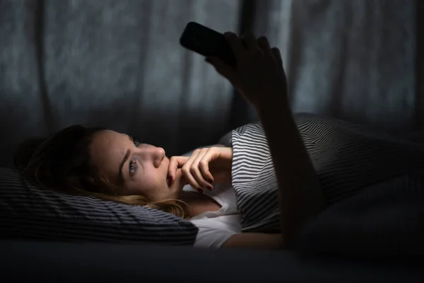 ベッドの中で若い女性は 疲れと疲れ 青い光が彼女の目を訓練し 彼女の概日リズムを台無しに電話を保持 — ストック写真