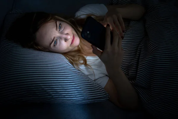 ベッドの中で若い女性は 疲れと疲れ 青い光が彼女の目を訓練し 彼女の概日リズムを台無しに電話を保持 — ストック写真