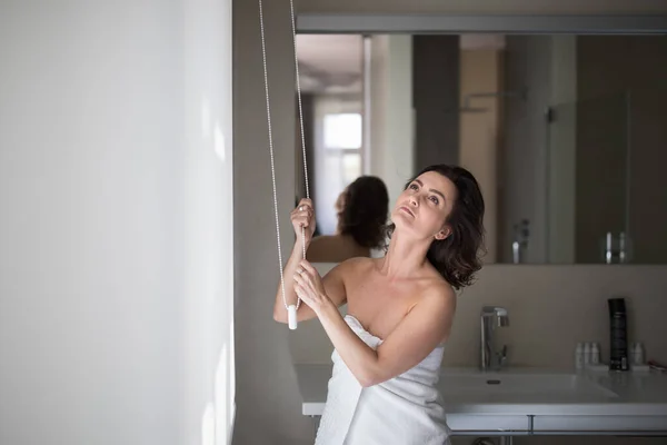現代のアパートで窓に白いローラーブラインドを開く中年の女性 — ストック写真