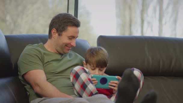 爸爸和儿子穿着睡衣坐在家里的沙发上 用手持设备玩电脑游戏 慢动作射击 — 图库视频影像