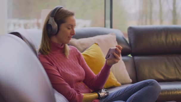自宅でソファに座って携帯電話から音楽ストリーミングを聞いて無線ヘッドフォンを身に着けている女性 スローモーションで撮影 — ストック動画