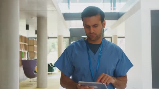 忙しい病院の建物を通って歩くデジタルタブレットを使用してスクラブを身に着けている男性医師 スローモーションで撮影 — ストック動画