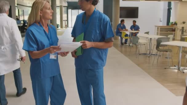 カメラトラックに沿って忙しい病院の廊下に男性と女性の医師は 患者のメモを議論スクラブを身に着けています スローモーションで撮影 — ストック動画