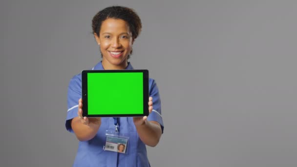 笑顔の女性看護師のスタジオポートレートは 灰色の背景に空白の緑の画面と均一な保持デジタルタブレットを身に着けています スローモーションで撮影 — ストック動画