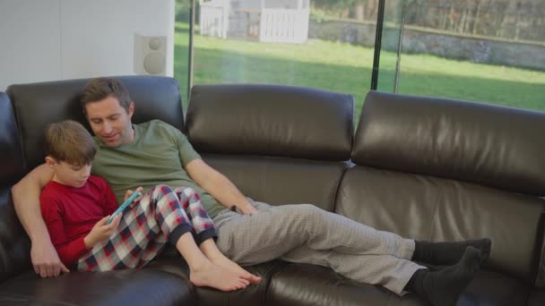 爸爸和儿子穿着睡衣躺在家里的沙发上 用手持设备玩电脑游戏 慢动作射击 — 图库视频影像