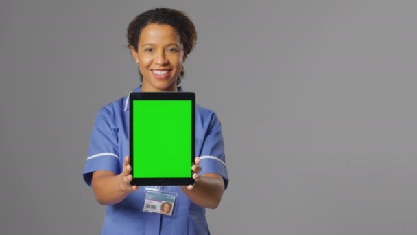 笑顔の女性看護師のスタジオポートレートは 灰色の背景に空白の緑の画面と均一な保持デジタルタブレットを身に着けています スローモーションで撮影 — ストック動画