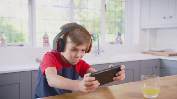 Boy Bär Trådlösa Hörlurar Hemma Köket Spelar Spel Mobiltelefon Och — Stockvideo