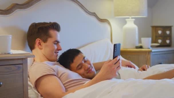 同じセックスの男性カップルを愛しますベッドで家に横たわって男と一緒に携帯電話上のソーシャルメディアを見ながらパートナー睡眠 遅い動きで撮影 — ストック動画