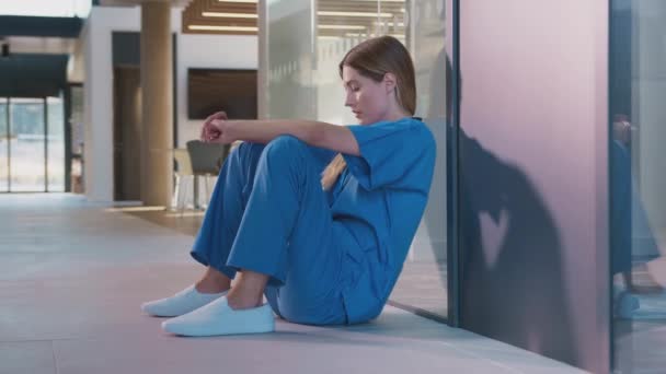 休憩中に病院の廊下の床に座っているスクラブの疲れと過労の女性医療従事者 スローモーションで撮影 — ストック動画