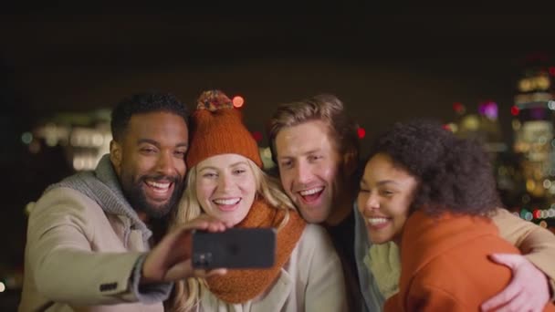 冬のコートやスカーフを身に着けている友人のグループは 背景に都市の照明と携帯電話で自撮りのためにポーズ スローモーションで撮影 — ストック動画