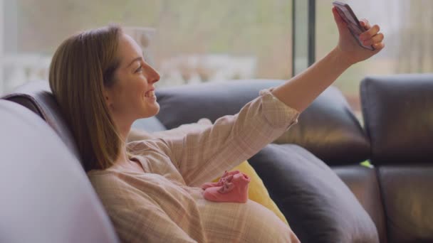 Pregnant Woman Sitting Sofa Home Taking Selfie Small Baby Shoes — Stock Video