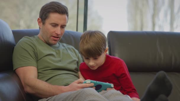 爸爸和儿子穿着睡衣坐在家里的沙发上 用手持设备玩电脑游戏 慢动作射击 — 图库视频影像