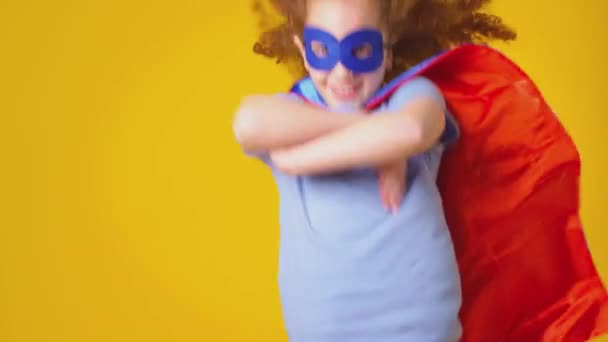 スタジオショットの女の子として身に着けている漫画の本スーパーヒーローを身に着けているマスクとケープふりをして飛ぶために黄色の背景 スローモーションで撮影 — ストック動画