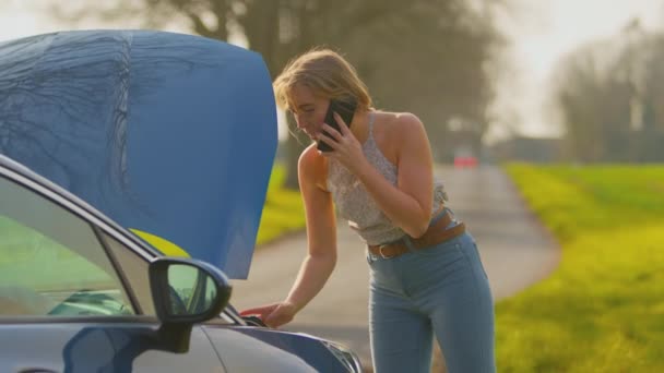 乡村道路上沮丧的女人站在被撞坏的汽车旁边 戴着便帽 用手机叫人帮忙 动作缓慢 — 图库视频影像