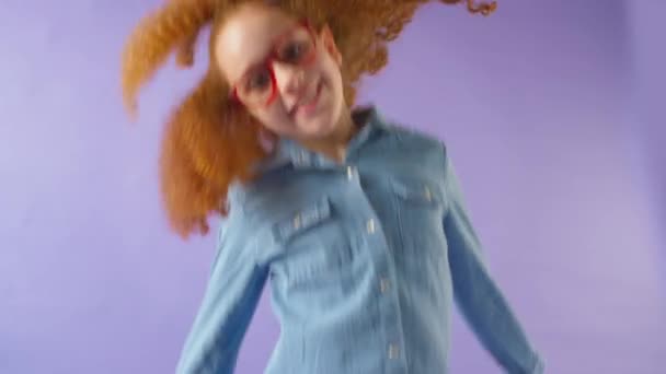 スタジオショットの笑顔の女の子とともに赤い髪と眼鏡ジャンプでフレームに紫のスタジオの背景 遅い動きで撮影 — ストック動画