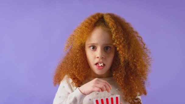 演播室里拍摄的红头发少女看恐怖片和吃爆米花的镜头 这些镜头都是她吓得在紫色演播室的背景下拍的 动作缓慢 — 图库视频影像