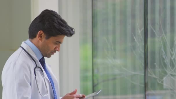 男性医師身に着けていますホワイトコート立ってで病院の廊下見ますデジタルタブレット 撮影でスローモーション — ストック動画