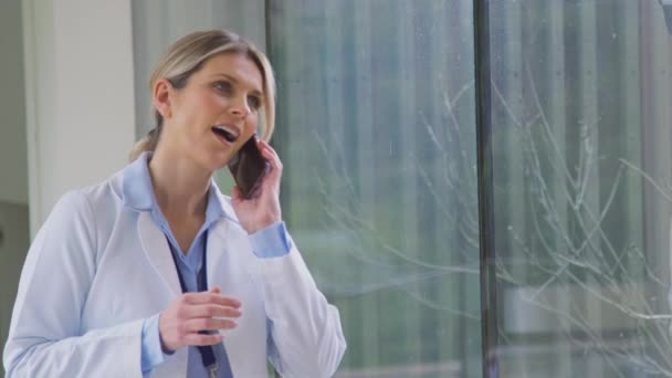 病院の廊下で携帯電話で話している白いコートを身に着けている女性医師 スローモーションで撮影 — ストック動画