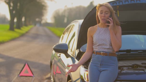 乡间道路上的危险警告标志 旁边站着一个女人 戴着便帽 站在被撞坏的汽车旁边 用慢镜头在手机上呼救 — 图库视频影像