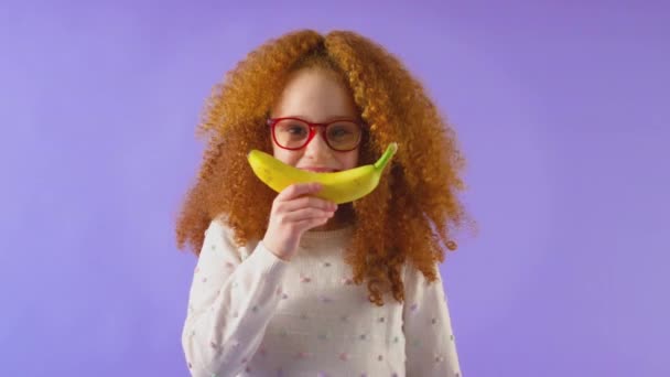 摄影棚拍摄的红头发少女戴着眼镜 嘴角挂着香蕉 像在紫色摄影棚背景下微笑 动作缓慢 — 图库视频影像