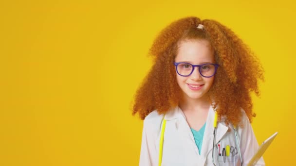 スタジオショットの女の子として服を着た医師や外科医とともにデジタルタブレット身に着けていますホワイトコートとともに聴診器で黄色の背景 遅い動きで撮影 — ストック動画