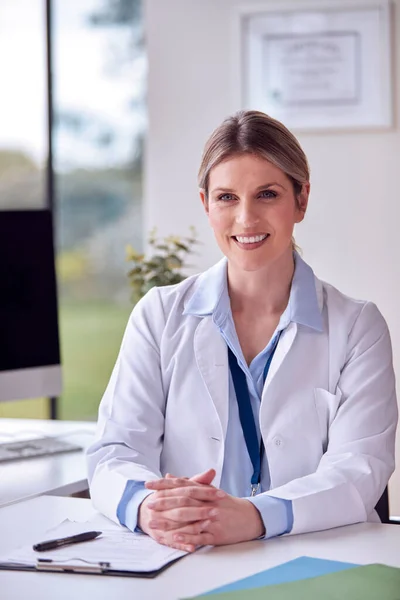 Portrait Of Smiling Female Doctor Or GP Wearing White Coat In Office Sitting At Desk