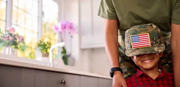 American Army Mother In Uniform Home On Leave With Son Wearing Cap In Family Kitchen