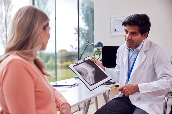 Doctor Wearing White Coat In Office With Mature Female Patient Looking At Medical X-Ray Or Scan