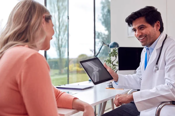 Doctor Wearing White Coat In Office Showing Mature Female Patient X-Ray Or Scan On Digital Tablet