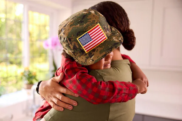 American Army Mother In Uniform Home On Leave Hugging Son Wearing Her Cap In Family Kitchen