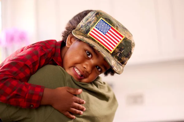 American Army Mother In Uniform Home On Leave Hugging Son Wearing Her Cap In Family Kitchen