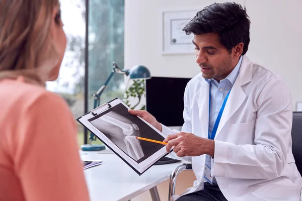 Doctor Wearing White Coat In Office With Mature Female Patient Looking At Medical X-Ray Or Scan