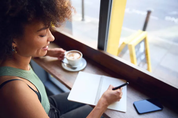 Female Customer In Coffee Shop Window Working Writing In Notebook