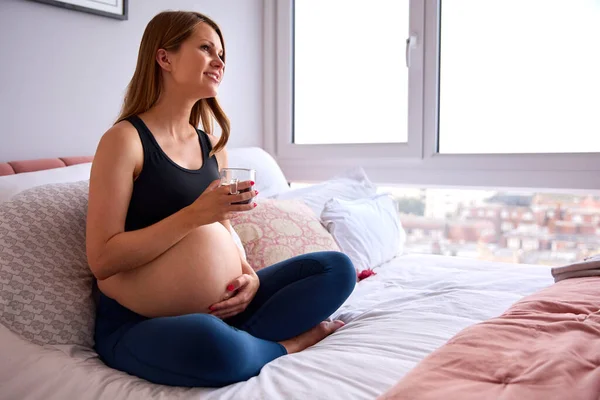 Embarazada Mujer Con Bebida Caliente Relajarse Cama Casa Sosteniendo Estómago —  Fotos de Stock