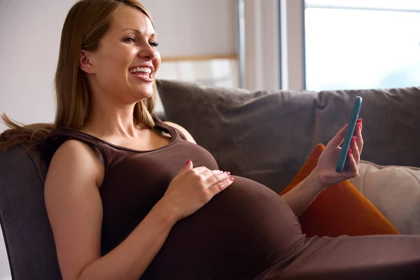 Mujer Embarazada Con Teléfono Móvil Relajante Sofá Casa Sosteniendo Estómago —  Fotos de Stock