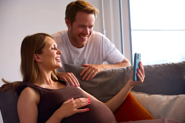 夫妇与怀孕妇女手握手机在沙发上轻松自在地持家胃 — 图库照片