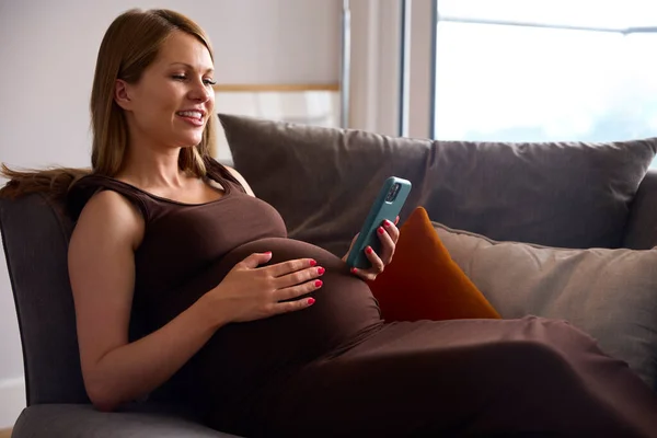 Těhotná Žena Mobilním Telefonem Relaxační Pohovce Doma Držení Břicha — Stock fotografie