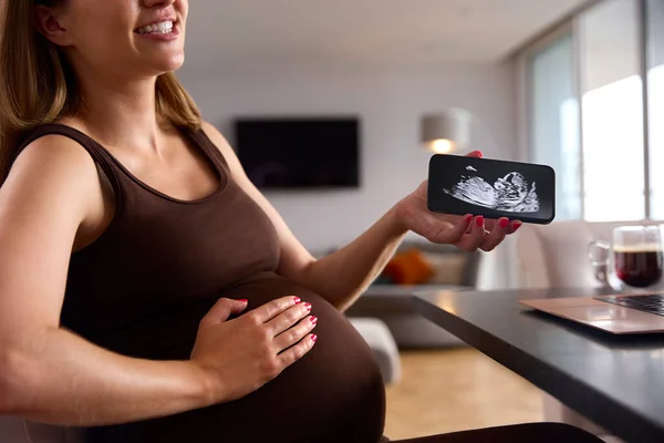 妊娠中の女性は赤ちゃんの超音波スキャンで携帯電話を見てノートパソコン上で自宅から働く — ストック写真