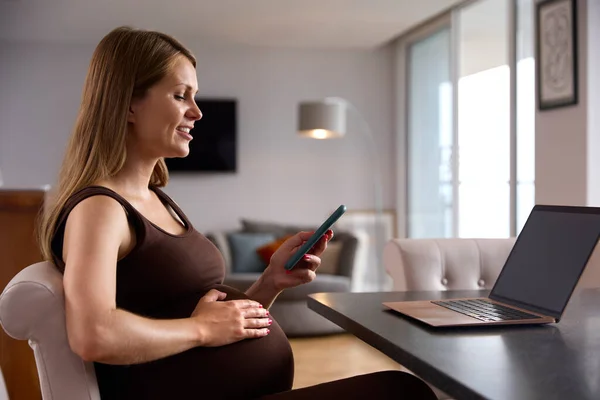 妊娠中の女性が自宅からノートパソコンで働くテーブルに座って電話を見て胃に触れる — ストック写真