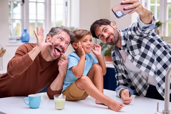 2人のお父さんと同じセックス家族カウンターに座っている息子とキッチンでセルフィーのために顔を引っ張る — ストック写真
