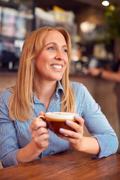 コーヒーショップのテーブル席に座る笑顔の女性が熱い飲み物を楽しむ — ストック写真