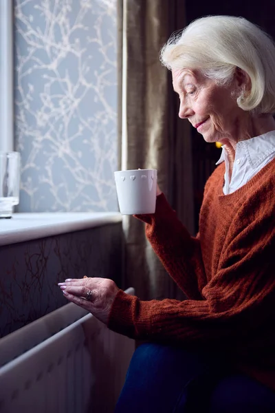 熱い飲み物を持つシニア女性は 生活エネルギー危機の費用で自宅でラジエーターで暖かくしようとしています — ストック写真