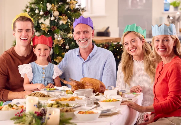 食事の前に紙の帽子をかぶって自宅でクリスマスを祝う世代の家族の肖像画 — ストック写真