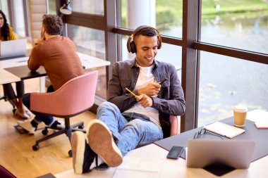 Young Businessman With Feet On Desk In Office Listening To Music On Wireless Headphones And Drumming clipart