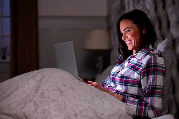 在家里微笑的女人在床上坐着 晚上用笔记本电脑 — 图库照片