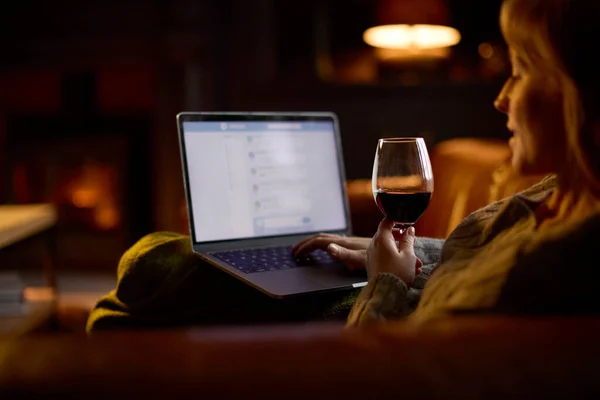 Woman At Home In Lounge Lying On Sofa With Cosy Fire Checking Emails On Laptop Holding Glass Of Wine