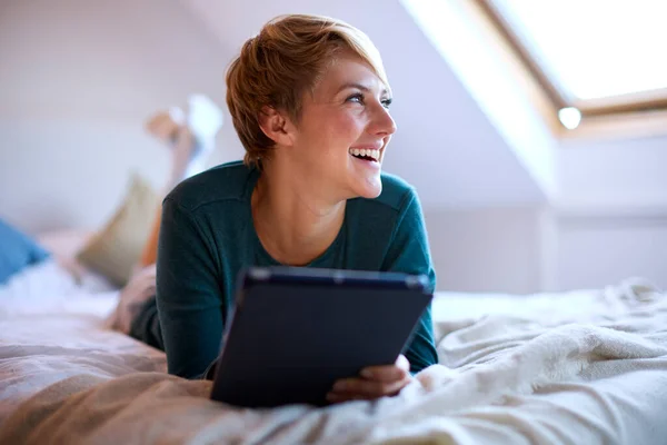若い笑顔の女性が自宅でパジャマを着てベッドの上で寝そべっているデジタルタブレットを見て — ストック写真