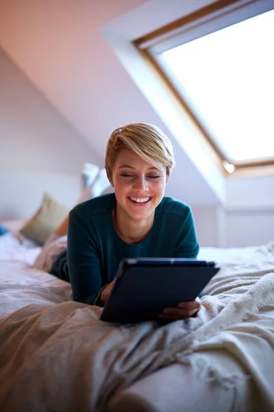 自宅でパジャマを着てベッドに寝そべっている若い女性デジタルタブレットを見て — ストック写真