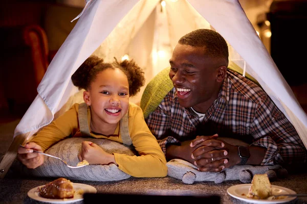 Father And Daughter At Home Lying In Indoor Tent Or Camp With Digital Tablet Eating Cake Together